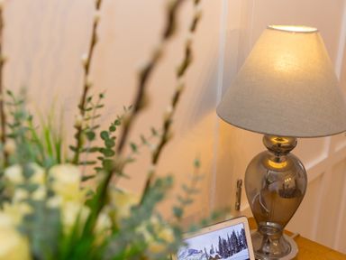 A table lamp with a beige shade next to a laptop on a wooden surface with flowers in the foreground at Captain's Walk in Newquay