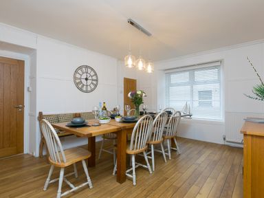 A dining room with a wooden table, six chairs, a bench, pendant lights, a window with blinds, and a wall clock at Captain's Walk in Newquay