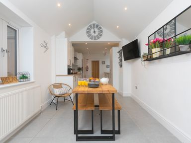 A dining area with a wooden table and benches next to a kitchen with white cabinets at Captains Walk in Newquay