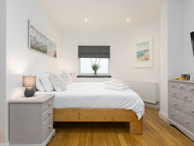 A bedroom with a wooden bed and white linens a nightstand with a lamp a window with a gray blind and flowers on the sill at Captain's Walk in Newquay