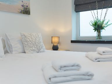 A bedroom with a bed with white linens and pillows a bedside table with a lamp and a window with a vase of flowers at Captain's Walk in Newquay