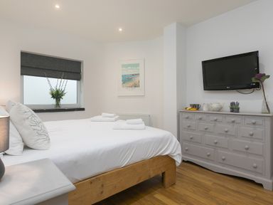 A bedroom with a wooden bed a gray dresser with a television mounted above and a window with a flower vase at Captain's Walk in Newquay