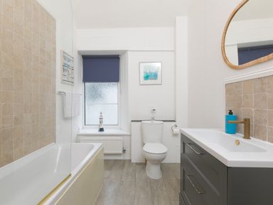 A bathroom with a bathtub tiled wall a toilet a sink with cabinet and a window with blue blind at Captain's Walk in Newquay