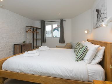 A bedroom with a wooden bed with white bedding and striped cushions next to a window with dark curtains and a wooden dresser with hangers at Captains Walk in Newquay