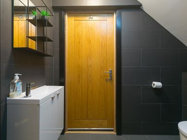 A bathroom with a wooden door a white sink cabinet with soap a toilet paper holder and a wall-mounted mirror with shelves at Captains Walk in Newquay