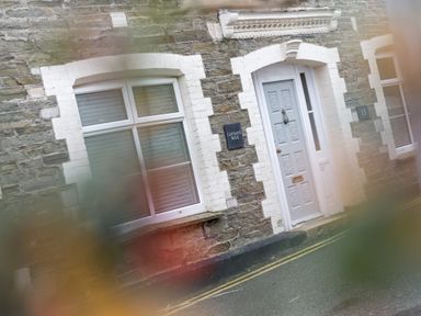 A stone house exterior with white window frames and a door with a sign reading Captains Walk in Newquay