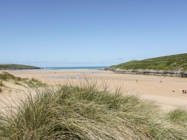 A sandy beach with grassy dunes and people walking near the shoreline at Captains Walk in Newquay