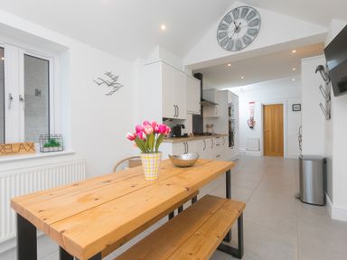 A kitchen with a wooden dining table and bench a vase with flowers a wall clock and white cabinets at Captain's Walk in Newquay