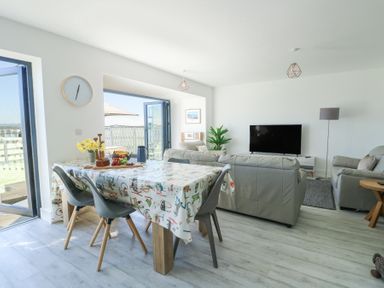 A living room and dining area with a dining table, chairs, sofas, a television, and an open door to an outdoor space at 6 Gwel Yr Wyddfa Llanfaelog near Rhosneigr