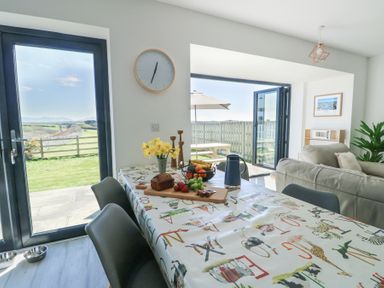 A dining area with a table covered by an alphabet tablecloth and fruit with a sofa and outdoor patio at 6 Gwel Yr Wyddfa Llanfaelog near Rhosneigr