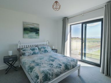 A bedroom with a double bed and two bedside tables next to a glass sliding door with curtains at 6 Gwel Yr Wyddfa in Llanfaelog near Rhosneigr