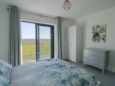 A bedroom with a bed, wardrobe, chest of drawers, and sliding glass doors showing a view of fields at 6 Gwel Yr Wyddfa in Llanfaelog near Rhosneigr