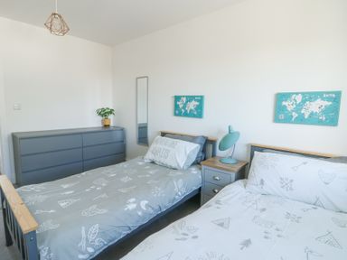 A bedroom with two single beds a gray chest of drawers a plant a turquoise lamp and world map pictures at 6 Gwel Yr Wyddfa Llanfaelog near Rhosneigr