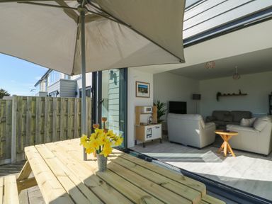 A wooden picnic table with yellow flowers and a patio umbrella next to sliding glass doors leading to a living room with sofas and a television at 6 Gwel Yr Wyddfa in Llanfaelog near Rhosneigr