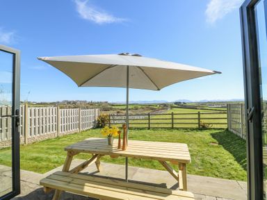 A wooden picnic table with a large umbrella in a fenced backyard overlooking fields and mountains at 6 Gwel Yr Wyddfa in Llanfaelog near Rhosneigr