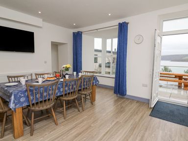 A dining room with a wooden table covered by a blue tablecloth and chairs set near a window and an open door showing an outdoor seating area at Fronwig in New Quay