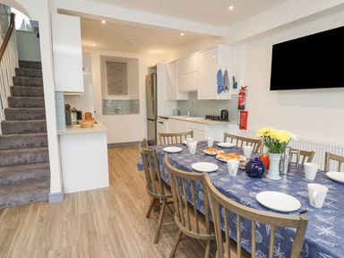 A dining area with a table set for a meal and a kitchen with white cabinets and appliances at Fronwig in New Quay
