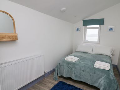 A small bedroom with a double bed covered with a green blanket and two white towels near a window with a green blind at Fronwig in New Quay