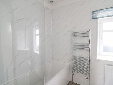 A bathroom with a bathtub a chrome towel radiator a window with a fish-patterned blind and marble-patterned walls at Fronwig in New Quay
