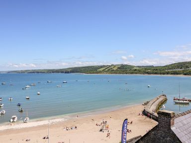 A beach with people, boats in the water, a pier, and grassy hills in the background at Fronwig in New Quay
