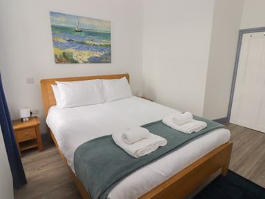 A bedroom with a wooden bed frame white bedding folded towels and a seascape painting on the wall at Fronwig in New Quay