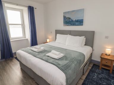 A bedroom with a double bed with white bedding and grey blanket beside a window with blue curtains at Fronwig in New Quay