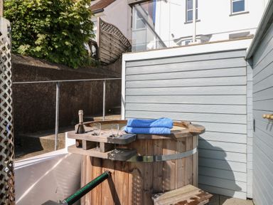 An outdoor wooden hot tub with a bottle of champagne and two glasses and blue towels on the edge at Fronwig in New Quay