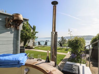 An outdoor seating area with a wooden hot tub a bottle of champagne two glasses and chairs overlooking a body of water at Fronwig New Quay