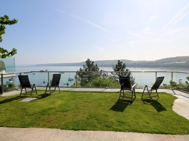 A grassy patio with four chairs facing a glass railing overlooking boats on water and hills at Fronwig in New Quay