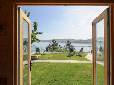 A garden with four chairs on grass overlooking water and hills through open wooden doors at Fronwig in New Quay