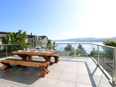 An outdoor patio with a wooden picnic table and benches set for dining overlooking a body of water with hills in the background at Fronwig in New Quay