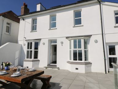 A patio area with a wooden dining table set with plates and glasses outside a white house with bay windows at Fronwig in New Quay