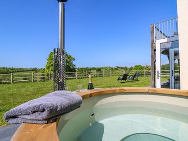 An outdoor hot tub with a towel a bottle of champagne and glasses on the edge and lawn chairs on grass at Yr Adfa in Llangeitho near Tregaron