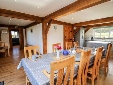A dining area with a table covered in a polka dot tablecloth and wooden chairs in a room with wooden beams and a kitchen in the background at Yr Adfa in Llangeitho near Tregaron