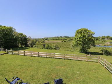A fenced grassy garden with outdoor chairs and trees overlooking fields and a pond at Yr Adfa in Llangeitho near Tregaron