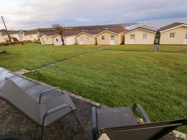 Two outdoor chairs on a patio facing a lawn with pathways and small houses beyond at Chalet 22 in Port Eynon