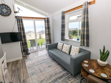 A living room with a gray sofa and patterned cushions a round wooden table with a plant a wall clock and a television at Chalet 22 in Port Eynon
