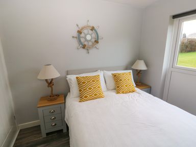A bedroom with a white bed and yellow patterned pillows two bedside tables with lamps and a window showing a green lawn at Chalet 22 in Port Eynon