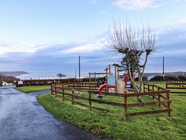 A fenced playground with slide and seesaw near a road and sea at Chalet 22 in Port Eynon