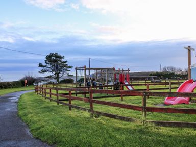 An outdoor playground with swings and slides surrounded by wooden fences and a paved path at Chalet 22 in Port Eynon