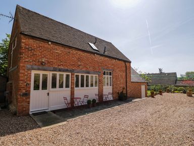 Exterior view of a brick building with white framed windows and doors with red tables and chairs outside at Home Farm Barn in Todenham near Moreton-In-Marsh
