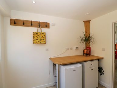 A corner with two small white refrigerators under a wooden counter with a potted plant on top and a wooden coat rack with a bag hanging at Home Farm Barn Todenham near Moreton-In-Marsh