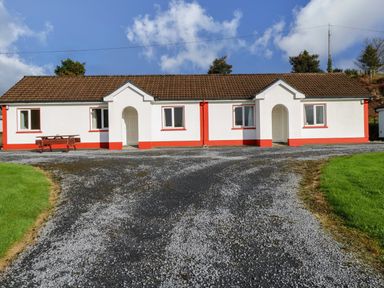 A bungalow with white walls and red trims on a gravel driveway with green grass on either side at Robins Roost 2 in Cornamona County Galway