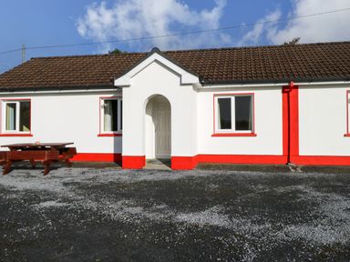 A white bungalow with red trim and a brown tiled roof with a wooden picnic table in front at Robins Roost 2 in Cornamona County Galway