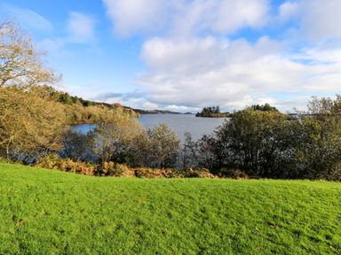 A grassy area overlooking a lake surrounded by trees under a partly cloudy sky at Robin's Roost 2 in Cornamona County Galway