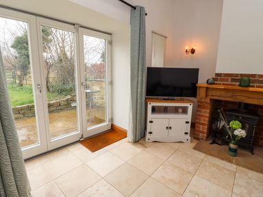 A room with tiled floor glass doors leading outside a television on a white stand and a brick fireplace at Christon Well Cottage in Christon Bank near Embleton