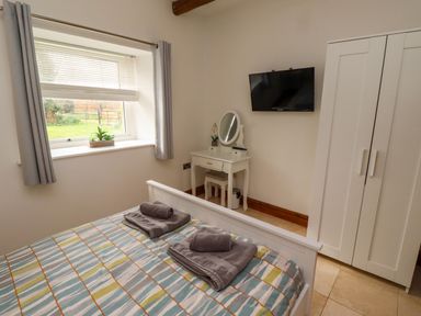 A bedroom with a bed, window with curtains, dressing table, wall-mounted TV, and wardrobe at Christon Well Cottage Christon Bank near Embleton