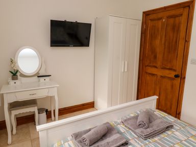 A bedroom with a bed with towels, a white vanity table with a mirror, a white wardrobe, a mounted TV, and a wooden door at Christon Well Cottage in Christon Bank near Embleton
