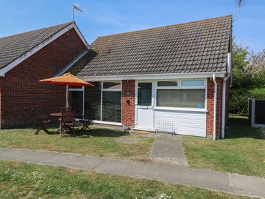A single-story brick house with a tiled roof a white door and windows a patio table with chairs and an orange umbrella on the lawn at 1 Kessingland Cottages in Kessingland