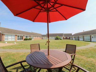 An outdoor wooden table with chairs and a red umbrella in a grassy courtyard surrounded by single-story buildings at 1 Kessingland Cottages in Kessingland